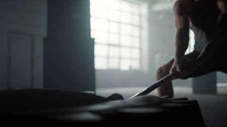 Closeup strong man hitting huge wheel with hammer in sport club. Professional bodybuilder silhouette...