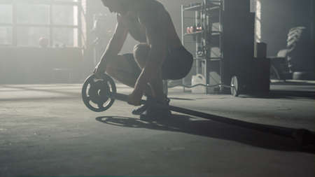 Closeup silhouette athletic man legs walking in dark fitness gym. Male bodybuilder preparing sports equipment for strength workout. Shirtless athlete putting heavy plates on barbell for trainingの写真素材