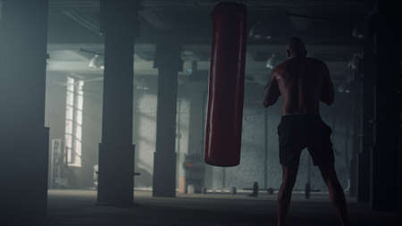 Muscular man boxing punch bag. Professional boxer training boxing in sports club. Powerful body sportsman punching in dark gym. Male athlete strength workout in boxing gloves. Sport motivation conceptの写真素材