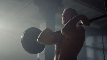 Male powerlifter lifting weights in dark fitness gym. Portrait powerful man holding heavy barbell on shoulders. Silhouette strong sportsman training body naked torso at strength workout in sport clubの写真素材