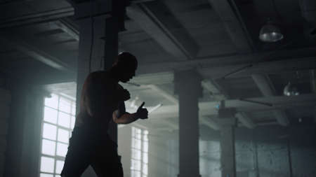 African American man training boxing in loft building. Male professional boxer fighting shadow in...