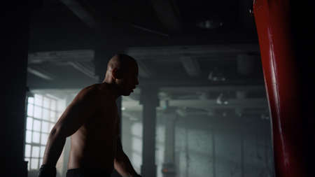 Tired boxer standing in sport club with punch bag. Exhausted athlete muscular sportsman resting at boxing training. African american powerful man body having break at strength workout in dark gymの写真素材