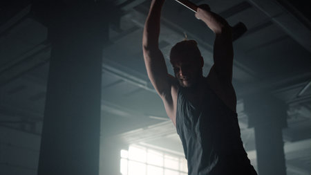 Aggressive man using hammer for strength exercise in sport club. Fitness guy hitting rubber tire during fitness training. Strong sportsman with sledgehammer doing intense workout in loft buildingの写真素材