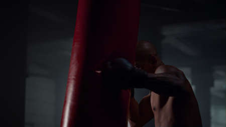Multiracial man boxing in fight club. Closeup male professional boxer hard training. Powerful sportsman kicking punching bag in dark gym. Close up strong athlete fighter naked torso strength workoutの写真素材