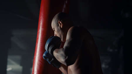 African american man box training in sport club. Close up professional boxer kicking punch bag at intensity workout. Powerful fighter exercising in dark gym. Muscular sportsman body. Speed strengthの写真素材