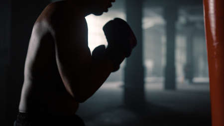 African american boxer boxing with punch bag. Athletic man actively moving during box training in gym. Strong male sportsman exercising in boxing gloves in sport club.の写真素材