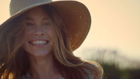 Happy woman face laughing in sunshine outdoor. Portrait of smiling girl walking away in green summer park. Beautiful lady in straw hat joking in illuminated sky background. Nice female face unfocusedの写真素材