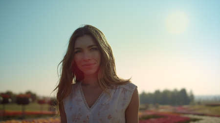 Beautiful woman face smiling camera in sunset background. Pretty girl straighten hair on flowers...