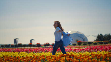 Back view of laughing girl running through tulip field with futuristic building in sunny day. Young woman moving fast in blooming garden in slow motion outdoors. Happy female feeling free in park.の写真素材