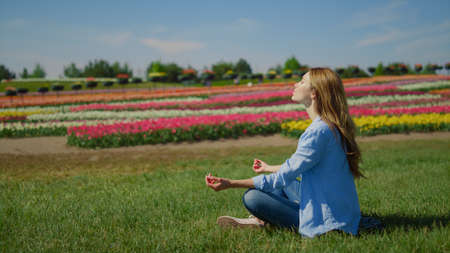 Young woman meditating on green grass in beautiful blooming park background. Hipster girl sitting in lotus in nature with flower landscape outdoor. Closeup woman profile making yoga in blossomed park.の写真素材
