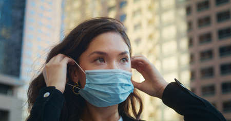 Closeup young asian woman using protective medical mask outdoors. Portrait of brunette covering face on cityscape. Beautiful girl care health safety practicing social distancing in pandemic.の写真素材