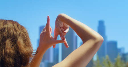 Unknown girl showing sign LA by fingers on blurred city background. Back view brunette making gestures name town Los Angeles close up. Woman standing in park enjoying beautiful summer cityscape.の写真素材