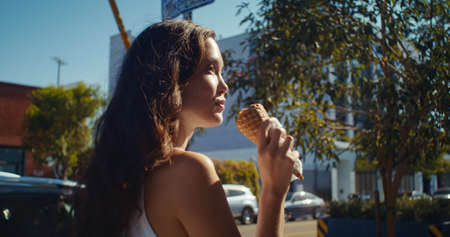 Happy asian girl licking ice cream walking city avenue side view. Attractive woman relaxing on summer weekend eating cold dessert outdoors. Dark hair lady tourist holding sweet street food closeup.の写真素材
