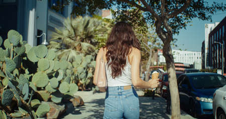 Unknown relaxing woman walking beautiful street holding ice cream back view. Stylish brunette enjoy...