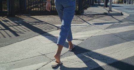 Unknown woman legs wearing stylish jeans walking crosswalk closeup. Unrecognizable girl crossing road in white comfortable shoes on sunny day. Female feet going across street on summer weekend.の写真素材
