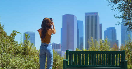 Unknown young woman standing in green park making photo skyscrapers on smartphone. Stylish girl taking picture blurred cityscape back view. Relaxed lady resting in public garden photographing far cityの写真素材