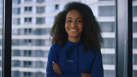Happy african american businesswoman looking camera in modern worklpace. Smiling female professional crossing hands in window office. Portrait of cute girl posing in company. Corporate people concept.の写真素材