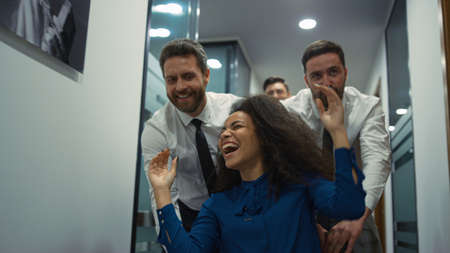 Multi ethnic coworkers team celebrating corporate success racing office chair in workplace. Diverse...