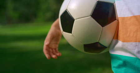 Close up soccer ball in hand young sportsman summer holiday. Unrecognized sporty boy holding soccerball on green grass park wearing colorful t-shirt at sunny day. Healthy active lifestyle concept.の写真素材