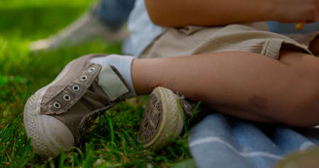 Unknown boy legs seat in gray sneakers on blue blanket close up. Unrecognizable little kid resting with family holding snacks on green park on weekend. Active leisure on summer nature concept.の写真素材