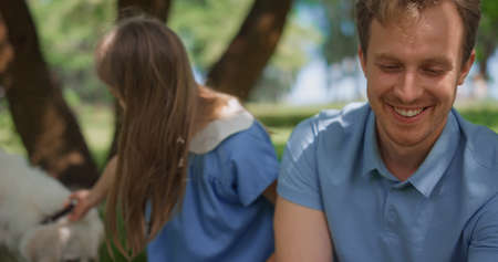Cheerful family rest on sunny nature using tablet closeup. Happy parents having fun with cute daughter playing with white dog in park on weekend. People enjoying digital technology on picnic outdoors.の写真素材