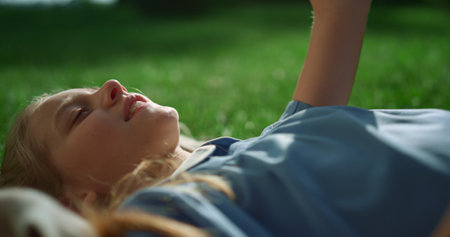Smiling blonde girl lying blanket on green field holding hand up closeup. Joyful kid enjoying golden sunlight in park. Child looking up at racket on weekend picnic alone. Summertime chill concept.の写真素材