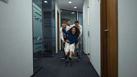 Multi ethnic coworkers team enjoy business success riding office chair in modern workplace. Smiling employees having fun celebrating friday in corporation company corridor. Corporate happy concept.の写真素材
