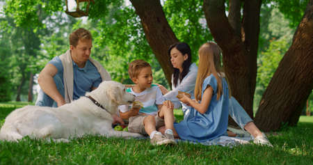 Happy family feeding white dog with two children on summer picnic. Little smiling boy give labrador...