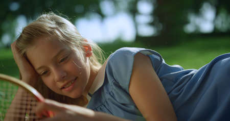 Little smiling girl lying on green lawn. Blond kid touch wooden racket net closeup. Cheerful child relaxing examining badminton equipment in park in golden sunlight. Summertime activities concept.の写真素材