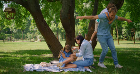 Little son have piggyback ride on happy father summer picnic. Agile man hold cute boy on back running near playing mother with daughter. Playful family have fun on beautiful nature in tree shadow.の写真素材