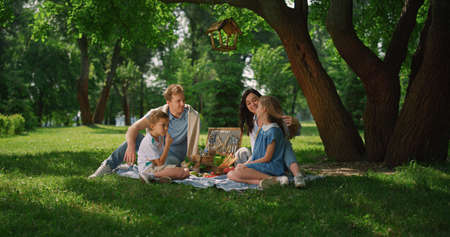 Happy two parent family enjoy picnic in summer park on holiday. Cheerful people lunching on green grass under tall tree. Resting family sitting white blanket eating snacks from basket outdoors.の写真素材