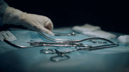 Table with surgical instruments in dark sterile operating room side view closeup. Unrecognized doctor assistant handing equipment help surgeon in emergency clinic ward. Modern medical tools concept.の写真素材