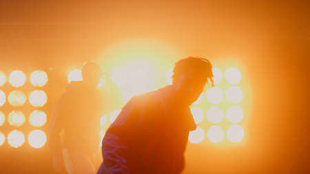 Dancing men silhouettes performing hip hop in bright spotlights closeup. Unknown agile guys moving energetically illuminated yellow nightclub lamps. Trendy team making freestyle show on stage indoorsの写真素材
