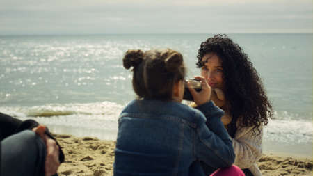 Lovely family playing photographer at beach. Mom child taking photos outdoors. Small girl photographing curly hair woman mother at ocean coast. Parent kid enjoy having fun on nature. Leisure concept.の写真素材