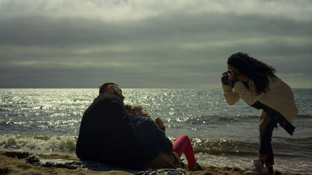 Happy family taking photos on ocean beach. People sitting posing camera by sea. Joyful parents child...