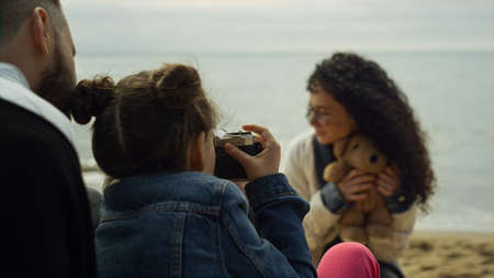 Family playing photo camera on sea beach. Parents child photographing on ocean coast. Father kid mother posing holding teddy bear on spring vacation. People relaxing on picnic. Childhood concept.の写真素材