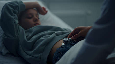 Little patient lying in clinic bed. Medical examination in hospital closeup. Unrecognized physician doctor listen girl stomach using stethoscope in emergency ward. Children healthcare system concept.の写真素材