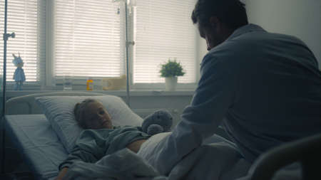 Pediatrician examining little patient in hospital ward. Girl lying in clinic bed. Professional male...