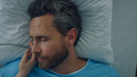 Ill man lying in hospital bed portrait. Patient recover after surgical operation. Unknown visitor female hand gently touching face expressing support. Loving wife take care of sick husband in clinic.の写真素材