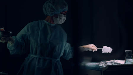 Female nurse assisting surgeon holding surgical instruments in clinic ward. Focused intern doctor...