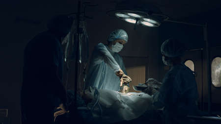 Confident surgeon performing operation in dark sterile clinic emergency room. Nurse assisting handing medical equipment to professional doctor in hospital ward. Healthcare workers teamwork concept.の写真素材