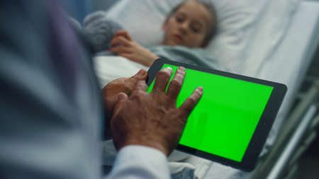 Doctor touching tablet green screen closeup. Sick girl lying in hospital bed. Unrecognized male medical workers hands using chroma key pad computer checking medical records. Modern healthcare system.の写真素材