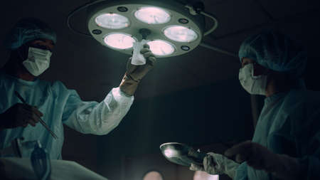 Doctor adjusting surgery lights in operating room. Hospital intensive care unit. African american man focused surgeon performing operation in sterile ward. Medical nurse assist holding instruments.の写真素材