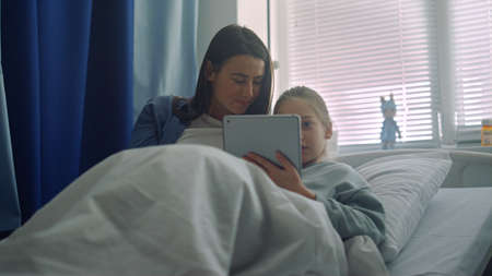 Smiling girl play pad with mother in ward. Supportive parent visiting sick kid. Joyful young mom hug...