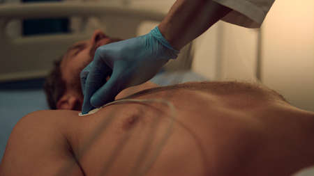 Cables on patient chest put by unknown cardiologist in modern hospital facility. Doctor specialist placing electrocardiogram ECG leads on skin in gloves. Healthcare professional using medical device.の写真素材