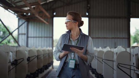Livestock worker walking animal husbandry. Professional breeder holding tablet. Focused woman farming engineer checking feedlots inspecting empty shed at countryside. Agricultural specialist conceptの写真素材