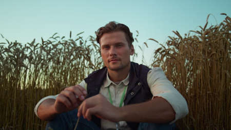 Portrait serious farmer posing at cultivated wheat field. Tired man rest alone. Thoughtful worker agronomist examining grain spikelets closeup. Successful agribusiness owner sit at evening farmland.の写真素材
