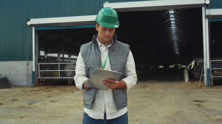 Livestock engineer making notes at modern cowshed building in protective helmet. Focused man farm...