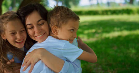 Happy kids cuddling smiling mother on summer picnic close up. Laughing blond siblings have fun sitting with mommy on green meadow. Woman enjoy embrace with children on weekend. Joyful family rest.の写真素材