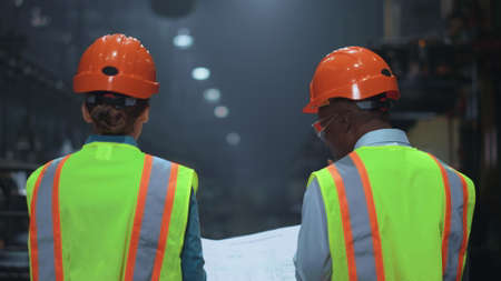 Professional multiethnic supervisors walking at special factory warehouse. Industrial mechanic colleagues discussing modern project work at manufacturing company. Partners wearing safety uniform.の写真素材